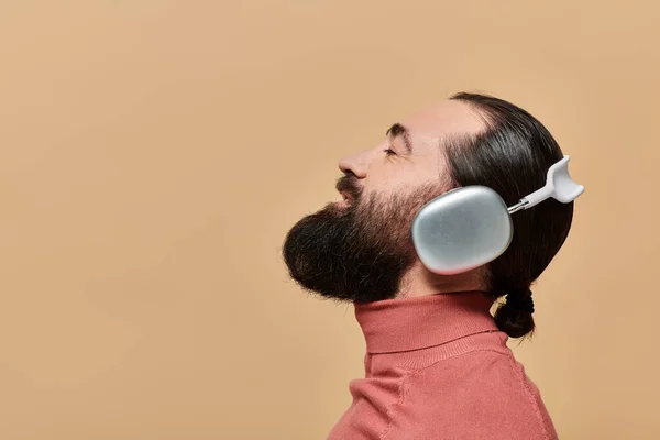 Side view, pleased bearded man in turtleneck jumper listening music in headphones, beige backdrop — Stock Photo