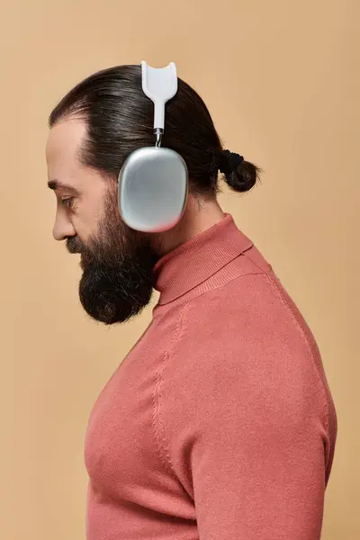 Side view, handsome bearded man in turtleneck jumper listening music in wireless headphones on beige — Stock Photo