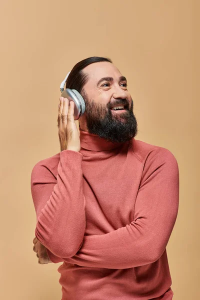Lächelnder und bärtiger Mann im Rollkragenpullover, der Musik in drahtlosen Kopfhörern hört, beige Hintergrund — Stockfoto