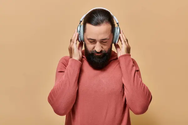 Schöner bärtiger Mann im Rollkragenpullover, der Musik in drahtlosen Kopfhörern auf beigem Hintergrund hört — Stockfoto