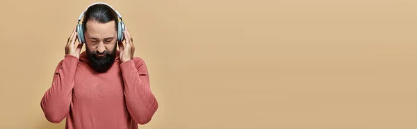 Handsome bearded man in turtleneck jumper listening music in wireless headphones on beige, banner — Stock Photo