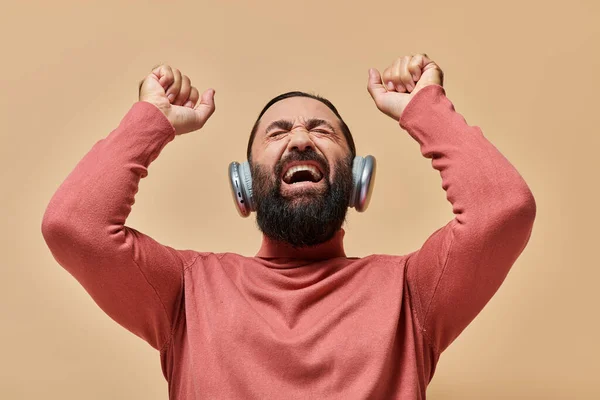 Uomo barbuto eccitato in maglione collo alto ascoltare musica in cuffie wireless, sfondo beige — Foto stock