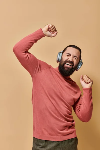 Excited bearded man in turtleneck jumper listening music in wireless headphones, beige background — Stock Photo