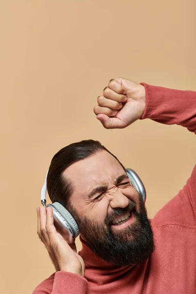 Взволнованный бородатый мужчина в водолазке джампер слушать музыку в беспроводных наушниках, бежевый фон — стоковое фото