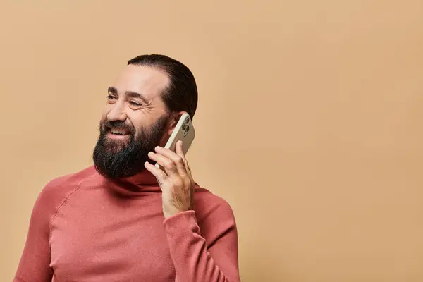 Portrait of cheerful bearded man in turtleneck jumper talking on smartphone on beige background — Stock Photo