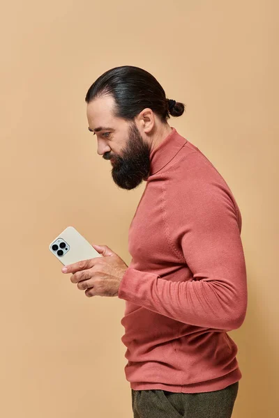 Portrait of bearded good looking man in turtleneck jumper holding smartphone on beige background — Stock Photo