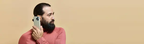 Портрет хорошого виглядає бородатого чоловіка в стрибку з горликом, що тримає смартфон на бежевому, банерному — стокове фото
