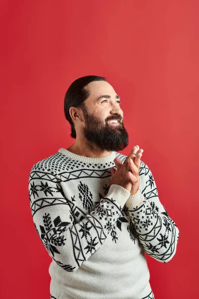 Hombre barbudo feliz en suéter de invierno con adorno sonriendo en el telón de fondo rojo, Feliz Navidad - foto de stock