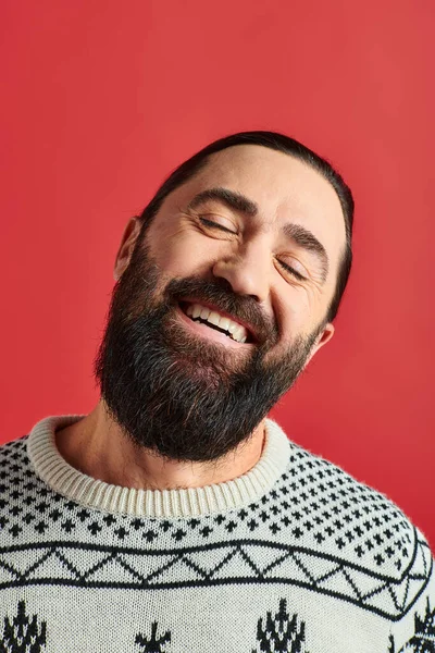 Hombre barbudo feliz en suéter de invierno con adorno sonriendo en el telón de fondo rojo, Feliz Navidad - foto de stock