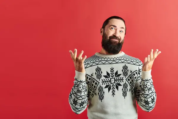 Confused bearded man in winter sweater with ornament smiling on red backdrop, Merry Christmas — Stock Photo