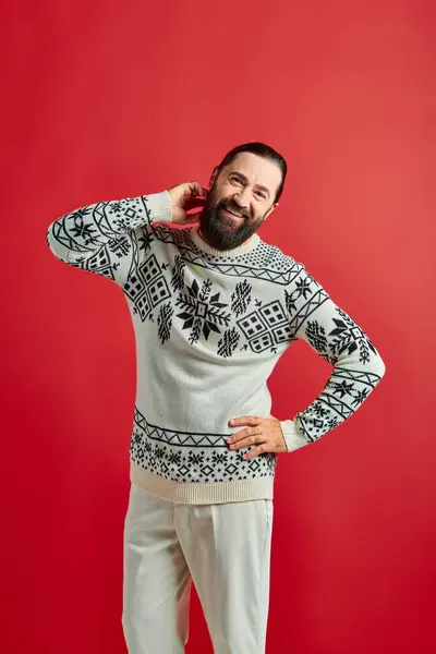 Hombre barbudo feliz en suéter de invierno posando con la mano en la cadera sobre el telón de fondo rojo, Feliz Navidad - foto de stock
