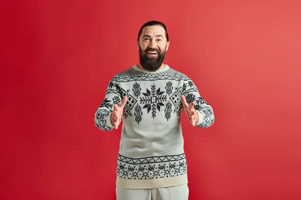 Positiver bärtiger Mann im Weihnachtspullover mit Ornament gestikulierend auf rotem Hintergrund, Winterurlaub — Stockfoto