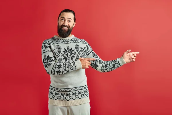 Hombre barbudo feliz en jersey de Navidad con adorno apuntando lejos en telón de fondo rojo, vacaciones de invierno - foto de stock