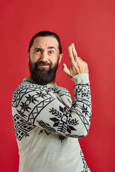 Happy bearded man in Christmas sweater showing gun gesture on red backdrop, winter holidays — Stock Photo