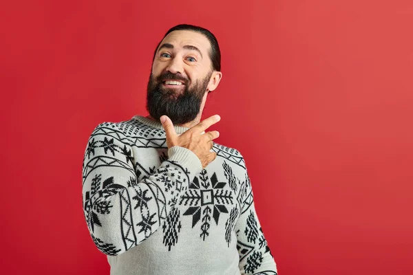 Hombre barbudo feliz en suéter de Navidad con adorno sonriendo en el telón de fondo rojo, vacaciones de invierno - foto de stock