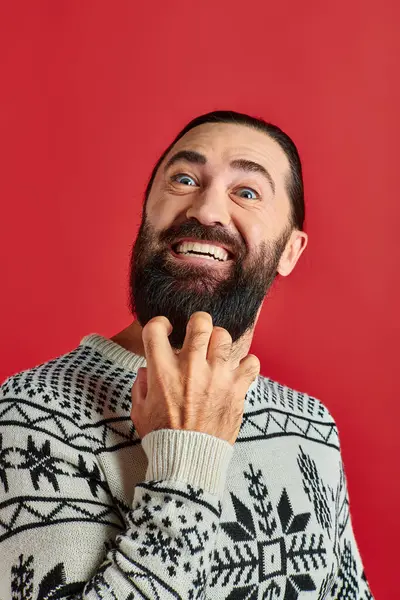 Hombre barbudo apasionado en suéter de Navidad con adorno sonriendo en el telón de fondo rojo, vacaciones de invierno - foto de stock