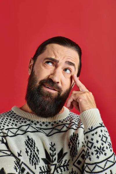 Hombre barbudo en jersey de Navidad con adorno pensando y apuntando a la cabeza sobre fondo rojo - foto de stock