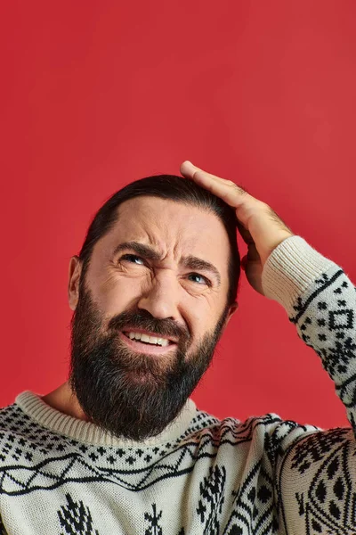 Бородатий чоловік на різдвяному светрі з орнаментом, що має головний біль і торкається голови на червоному тлі — стокове фото