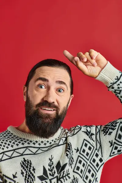 Happy bearded man in Christmas sweater with ornament pointing up on red backdrop, winter holidays — Stock Photo