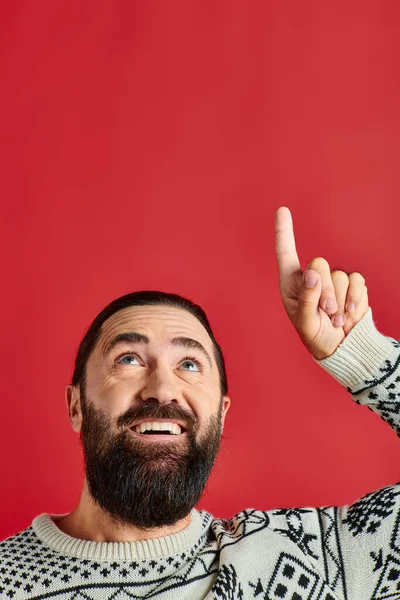 Happy bearded man in Christmas sweater with ornament pointing up on red backdrop, winter holidays — Stock Photo
