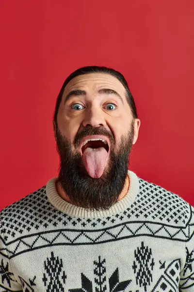 Hombre barbudo feliz en suéter con adorno lengua que sobresale sobre fondo rojo, Navidad - foto de stock