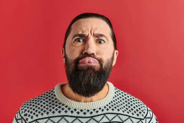 Bearded man in winter sweater with ornament sticking tongue out on red backdrop, Christmas — Stock Photo