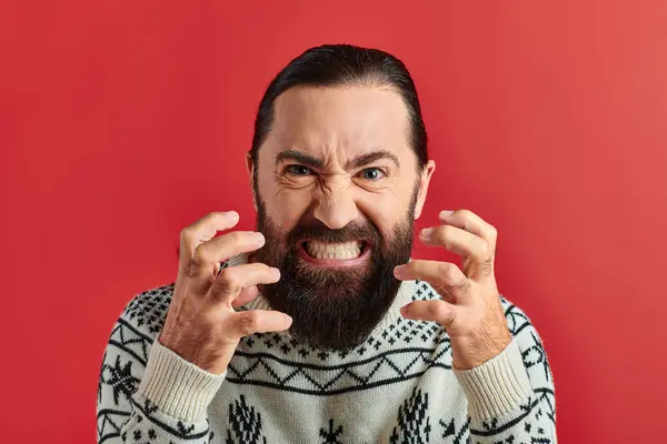 Сердитий бородатий чоловік в зимовому светрі з прикрасою, що посміхається і жестикулює на червоному тлі — стокове фото