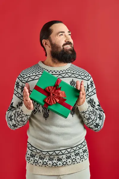 Heureux homme barbu en pull d'hiver avec ornement tenant cadeau de Noël sur fond rouge — Photo de stock