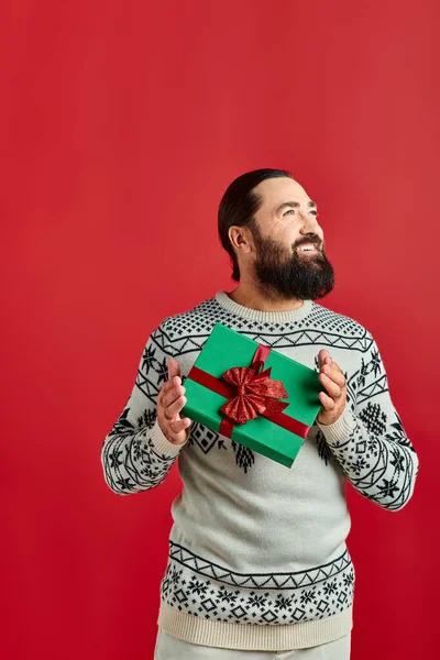 Щасливий бородатий чоловік в зимовому светрі з прикрасою, що тримає різдвяний подарунок на червоному тлі — стокове фото