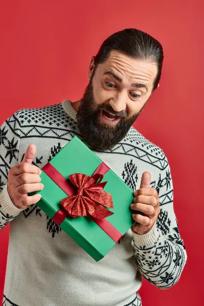Взволнованный бородатый мужчина в зимнем свитере с орнаментом держа рождественский подарок на красном фоне — стоковое фото