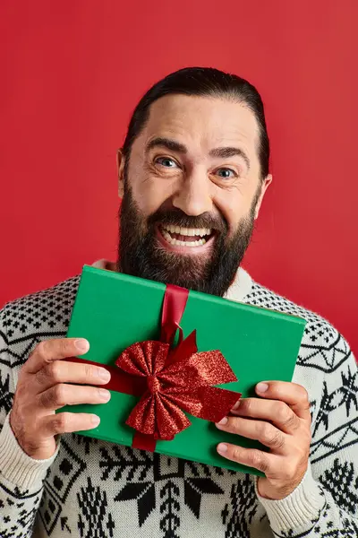 Excité homme barbu en pull d'hiver avec ornement tenant cadeau de Noël sur fond rouge — Photo de stock