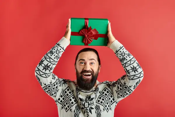Excité homme barbu en pull d'hiver avec ornement tenant cadeau de Noël sur fond rouge — Photo de stock
