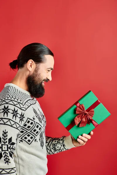 Вид сбоку, счастливый бородатый мужчина в зимнем свитере с рождественским подарком на красном — стоковое фото
