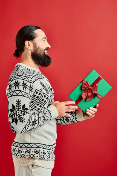 Вид сбоку, счастливый бородатый мужчина в зимнем свитере с рождественским подарком на красном — стоковое фото