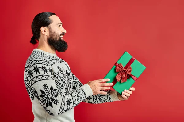 Side view, pleased bearded man in winter sweater with ornament holding Christmas present on red — Stock Photo
