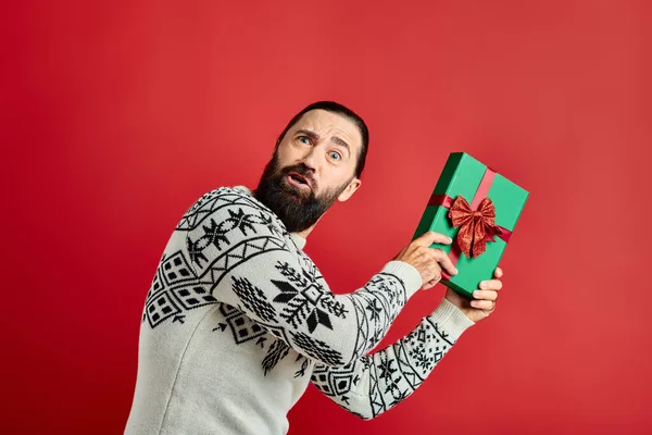 Homem barbudo confuso em camisola de inverno com ornamento segurando presente de Natal no fundo vermelho — Fotografia de Stock