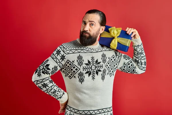 Bel homme barbu en pull d'hiver avec ornement tenant cadeau de Noël sur fond rouge — Photo de stock