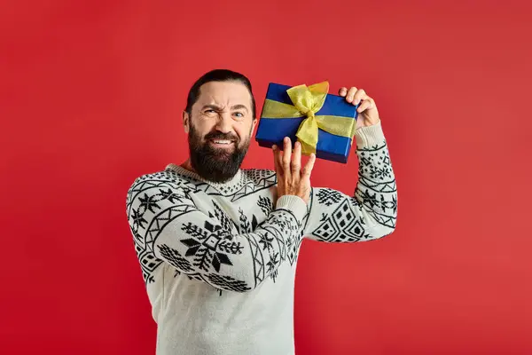 Missbilligte bärtige Mann im Winterpullover mit Ornament hält Weihnachtsgeschenk auf rotem Hintergrund — Stockfoto