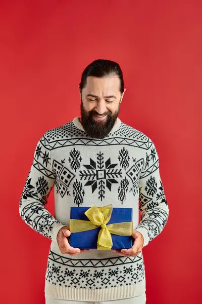 Heureux homme barbu en pull d'hiver avec ornement tenant cadeau de Noël sur fond rouge — Photo de stock