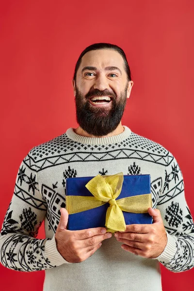Hombre barbudo complacido en suéter de invierno con adorno celebración regalo de Navidad sobre fondo rojo - foto de stock