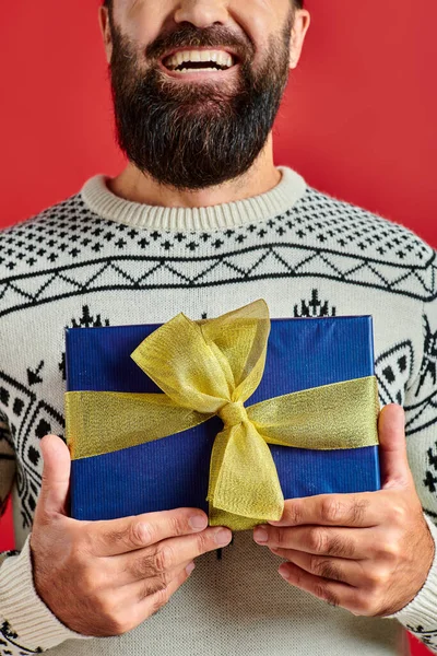 Обрезанный счастливый бородатый мужчина в зимнем свитере с орнаментом держа рождественский подарок на красном фоне — стоковое фото