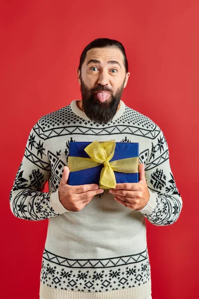 Funny bearded man in winter sweater with ornament holding Christmas gift and sticking tongue on red — Stock Photo