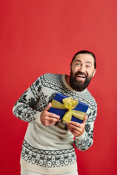 Joyeux homme barbu en pull d'hiver avec ornement tenant cadeau de Noël sur fond rouge — Photo de stock