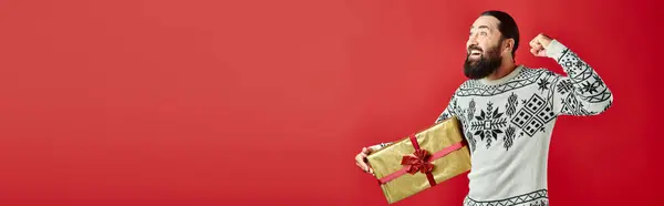 Aufgeregter bärtiger Mann im Winterpullover mit Ornament hält Weihnachtsgeschenk auf rot, Banner — Stockfoto