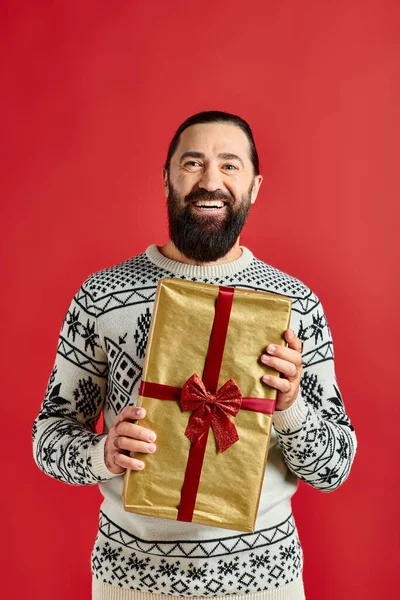 Bearded joyful man in winter sweater with ornament holding Christmas present on red background — Stock Photo