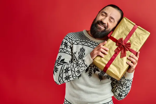 Homme joyeux barbu en pull d'hiver avec ornement tenant cadeau de Noël sur fond rouge — Photo de stock