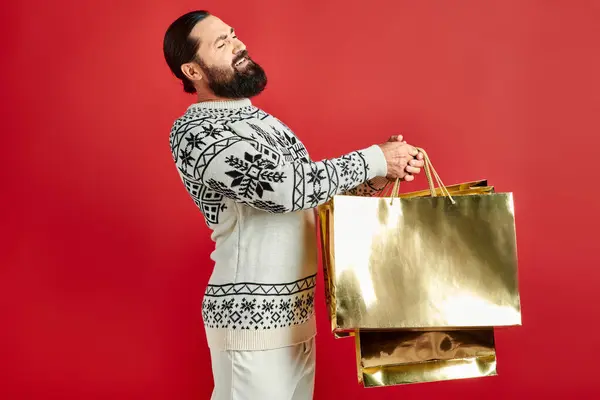 Недовольный бородатый мужчина в свитере с орнаментом держа сумки для покупок на красном фоне, Рождество — стоковое фото