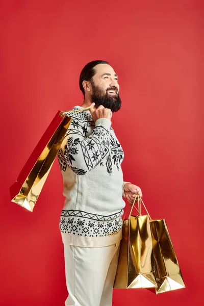 Glücklicher bärtiger Mann im Pullover mit Ornament, der Einkaufstüten auf rotem Hintergrund hält, Weihnachtsgeschenk — Stockfoto