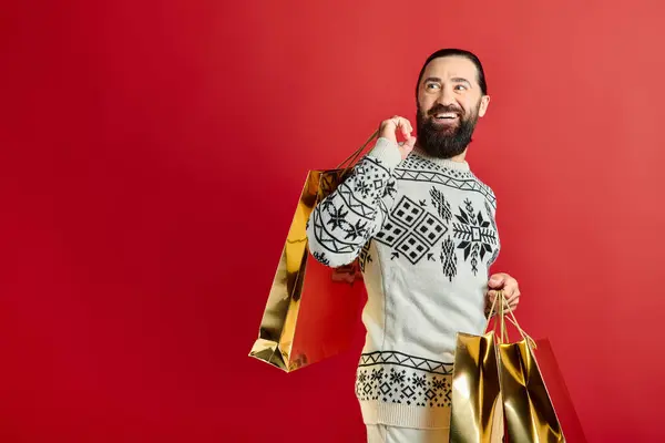 Изумленный бородатый мужчина в свитере с орнаментом держа сумки для покупок на красном фоне, рождественский подарок — стоковое фото