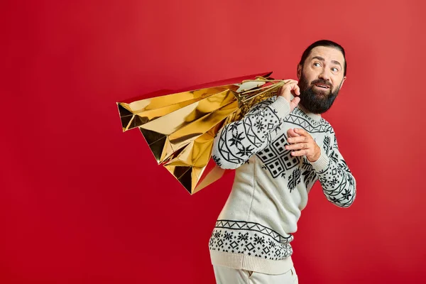 Joyeux homme barbu en pull avec ornement tenant des sacs à provisions sur fond rouge, cadeau de Noël — Photo de stock